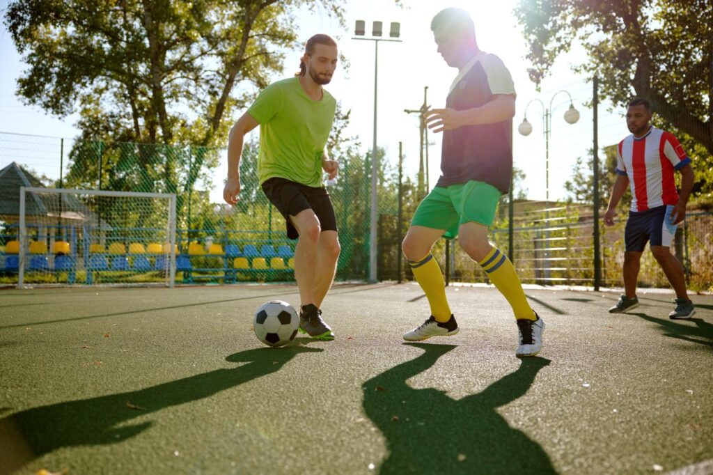 Homens jogando futebol em quadra com o sol batendo.