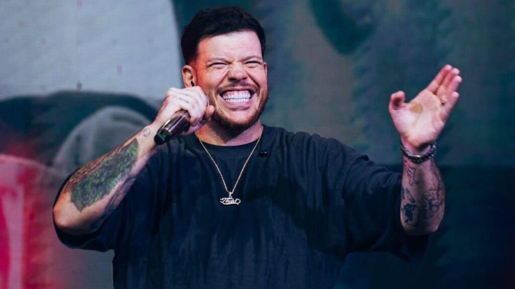Cantor Ferrugem. Homem branco, cabelo curto, barba, sorrindo no palco e cantando.