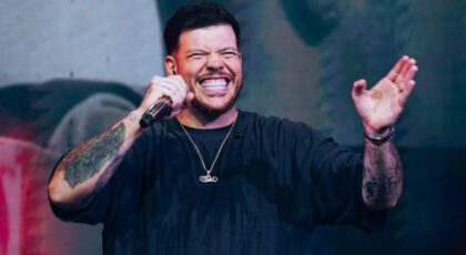 Cantor Ferrugem. Homem branco, cabelo curto, barba, sorrindo no palco e cantando.