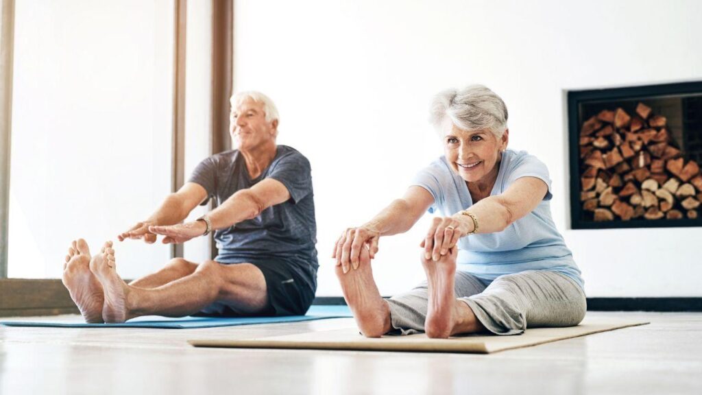 Imagem de dois idosos, um homem e uma mulher, fazendo alongamento no chão em tapetes de yoga