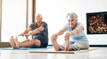 Imagem de dois idosos, um homem e uma mulher, fazendo alongamento no chão em tapetes de yoga