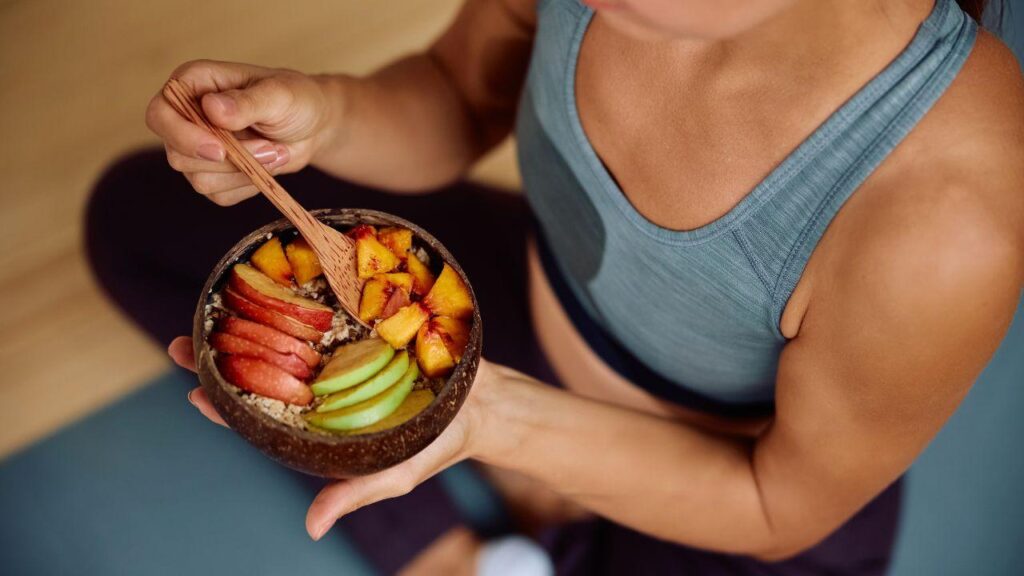 Imagem de uma mulher comendo seu lanche com roupa de academia, no pote que ela segura há frutas cortadas