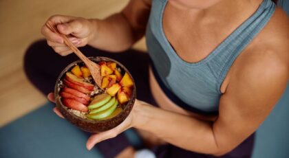 Imagem de uma mulher comendo seu lanche com roupa de academia, no pote que ela segura há frutas cortadas