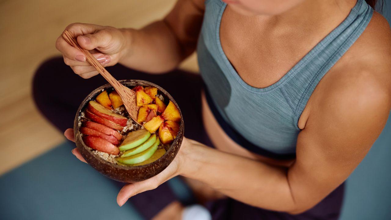 Imagem de uma mulher comendo seu lanche com roupa de academia, no pote que ela segura há frutas cortadas