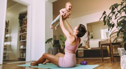Imagem de uma mulher sentada em um tapete de yoga, com roupa de treino, levantando a filha bebê no ar