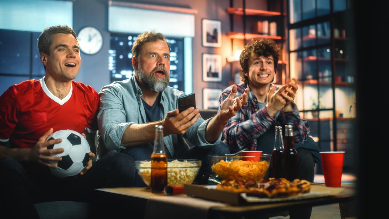 Três homens sentados no sofá assistindo a um jogo de futebol.