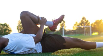 Foto mostra atleta de futebol deitado no campo do estádio de futebol com a mão na canela.