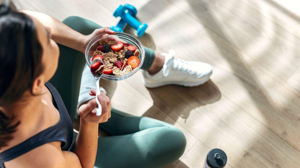 Imagem de uma mulher comendo cerais e frutas sentada no chão com halteres e uma garrafa de água ao lado