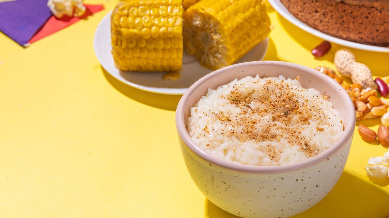 Imagem de arroz doce com canela em um pote, duas espigas de milho e um bolo redondo
