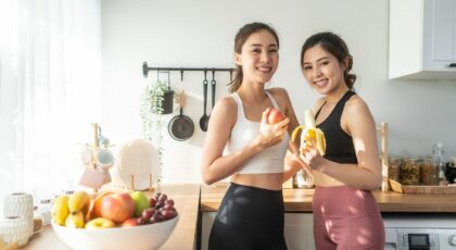 Imagem de duas mulheres jovens na cozinha, cada uma segurando uma fruta, com roupa de treino