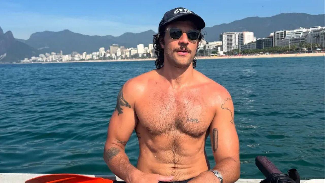 Chay Suede. Homem branco, cabelo curto castanho, sem camisa, abdômen definido, usando boné, barba e bigode ao ar livre.
