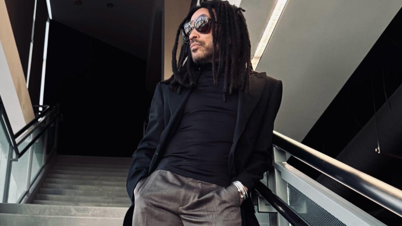 Foto mostra o cantor Lenny Kravitz de perfil, em pé na escada. Homem negro, com dread, óculos escuro, usando camisa marrom e jaqueta preta.