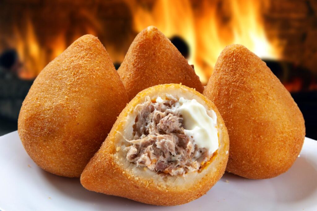 Foto mostra três coxinhas em cima de um prato em cima da mesa. Uma delas está partida mostrando o frango e catupiry.