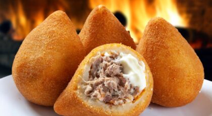 Foto mostra três coxinhas em cima de um prato em cima da mesa. Uma delas está partida mostrando o frango e catupiry.