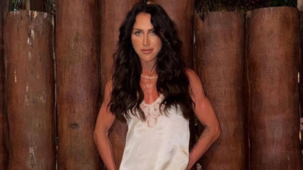 Foto mostra Aline Becker. Mulher branca, cabelo preto longo ondulado, usando vestido branco em um fundo de madeira.