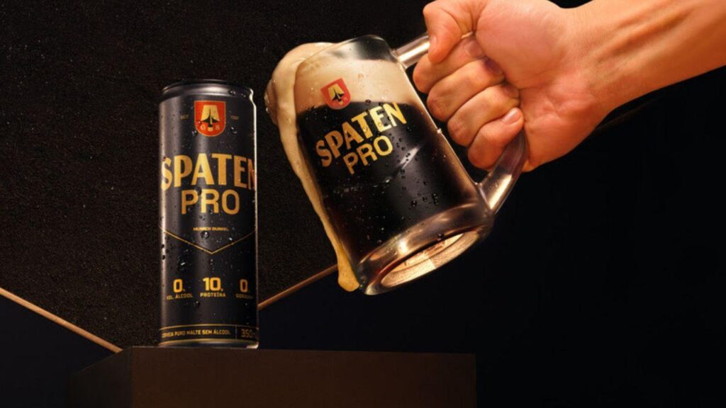 Foto mostra uma mão segurando a caneca de vidro de cerveja escura e brindando com a lata de cerveja.