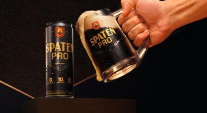 Foto mostra uma mão segurando a caneca de vidro de cerveja escura e brindando com a lata de cerveja.