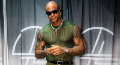 Foto mostra o cantor Léo Santana em pé, sorrindo para a câmera. Homem negro, careca, usa óculos escuro, camisa verde transparente e um colar pequeno.
