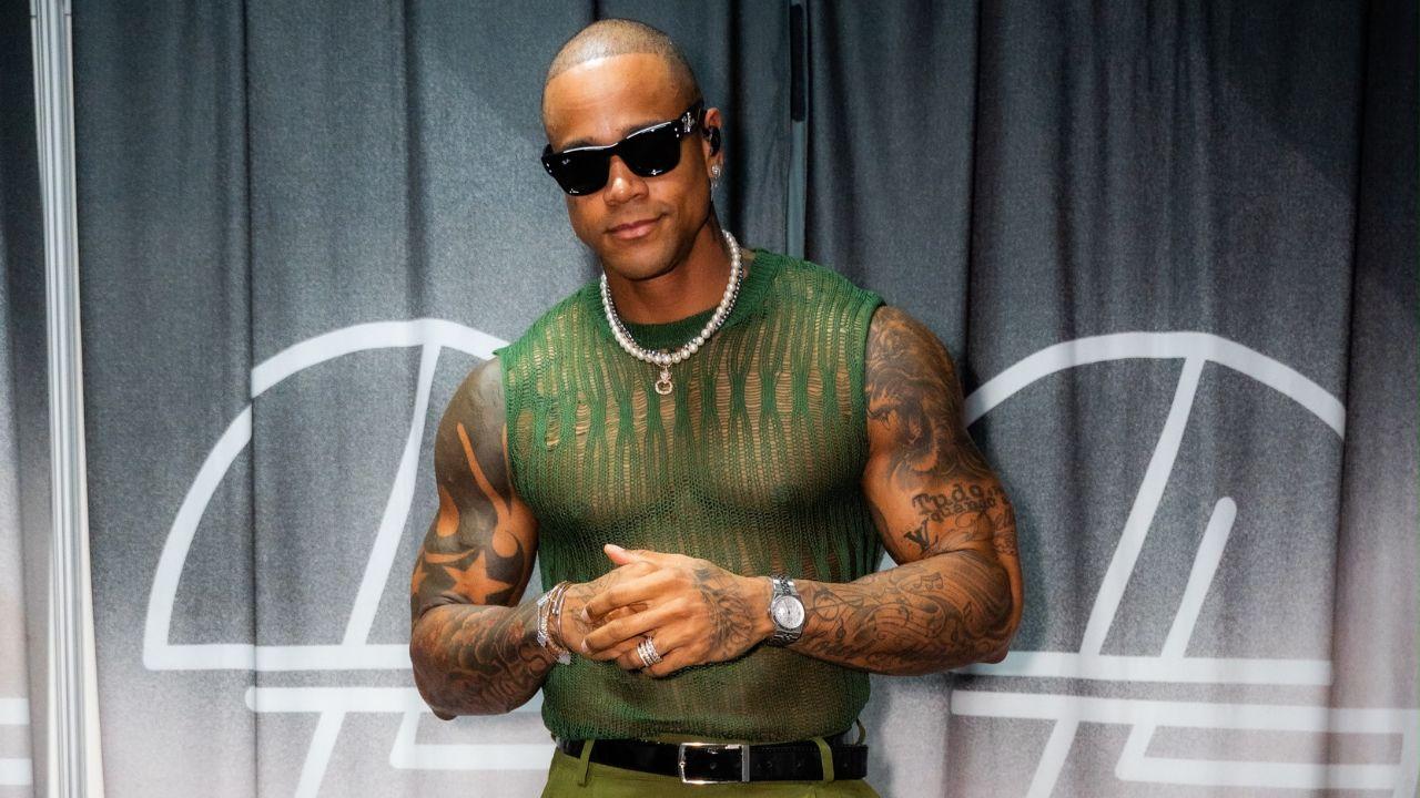 Foto mostra o cantor Léo Santana em pé, sorrindo para a câmera. Homem negro, careca, usa óculos escuro, camisa verde transparente e um colar pequeno.