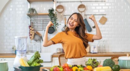 Mulher em pé na frente da bancada da cozinha com várias frutas e legumes.