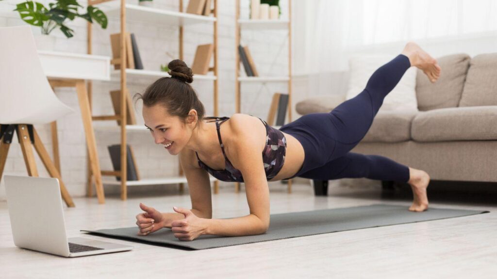 Imagem de uma mulher fazendo pilates na sala de casa em frente ao computador