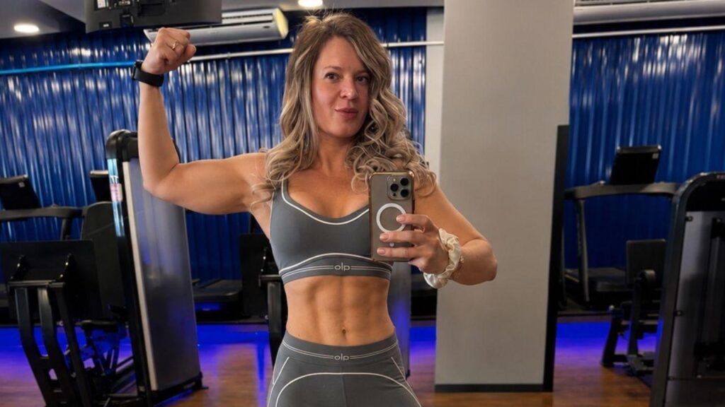 Joice Hasselmann. Mulher branca, cabelo loiro ondulado, com top cinza, tirando foto na frente do espelho com o biceps em destaque.