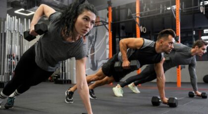 Mulher e homens jovens fazendo exercício na academia, levantando peso com um braço.