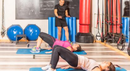 Duas pessoas fazendo o exercício chamado ponte em um colchonete com um professor assistindo.